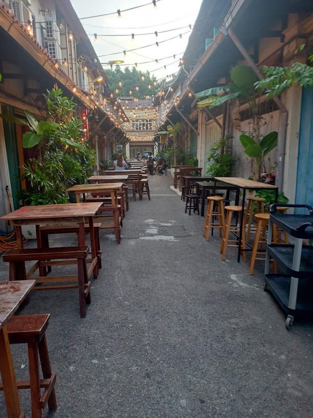 Outdoor seating along the alley beside Kafe Tuju, with wooden tables, stools and string lights overhead